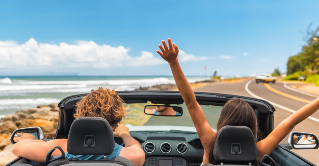 A couple sits in the driver's seat and passenger's seat of a convertible as we see their view of the road.