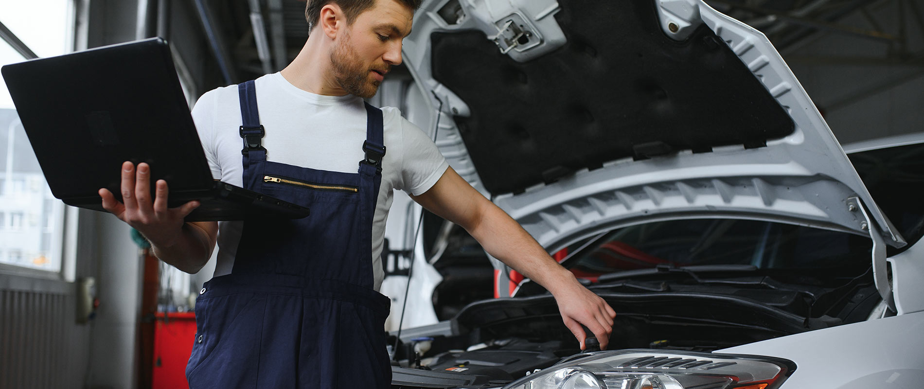 A mechanic examining the inside of a white car's hood while holding a laptop.