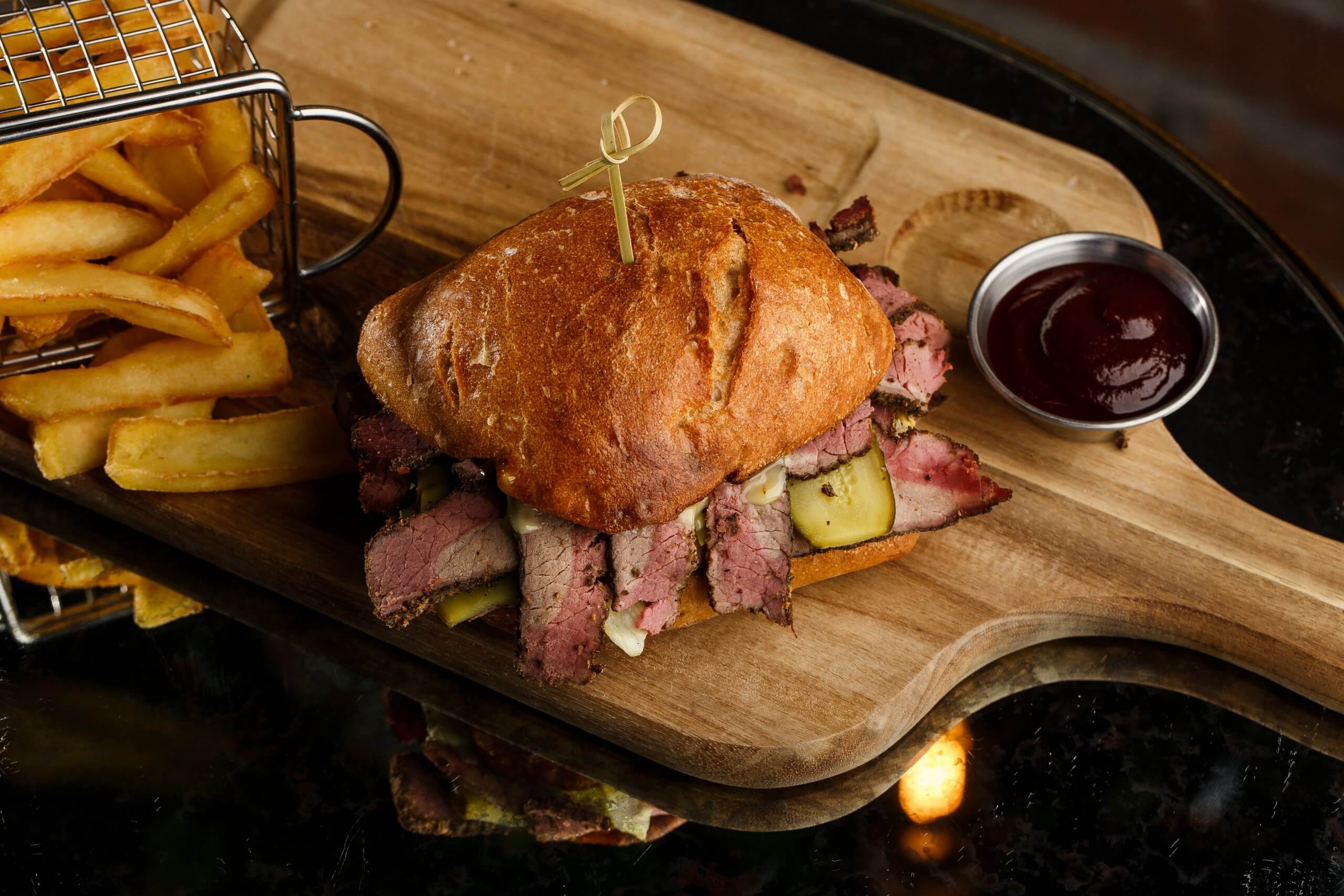 Pastrami sandwich on rye bread served with pickles and mustard sauce. Tasty sandwich with french fries potatoes on dark background.