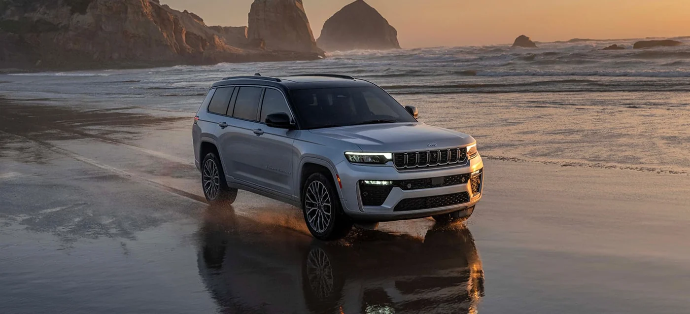 Silver Jeep Grand Cherokee L, the longer three-row body, parked on a wet beach at sunset