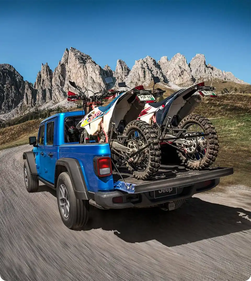 Blue Jeep Gladiator hauling two dirt bikes in its bed on a mountain road, tailgate down