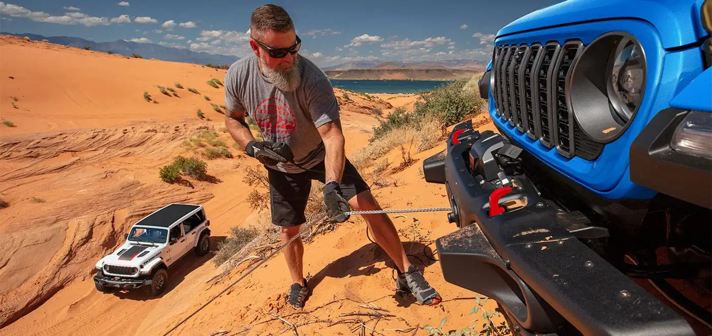 Two Jeep Wranglers on a sandy desert trail, one using a front winch line