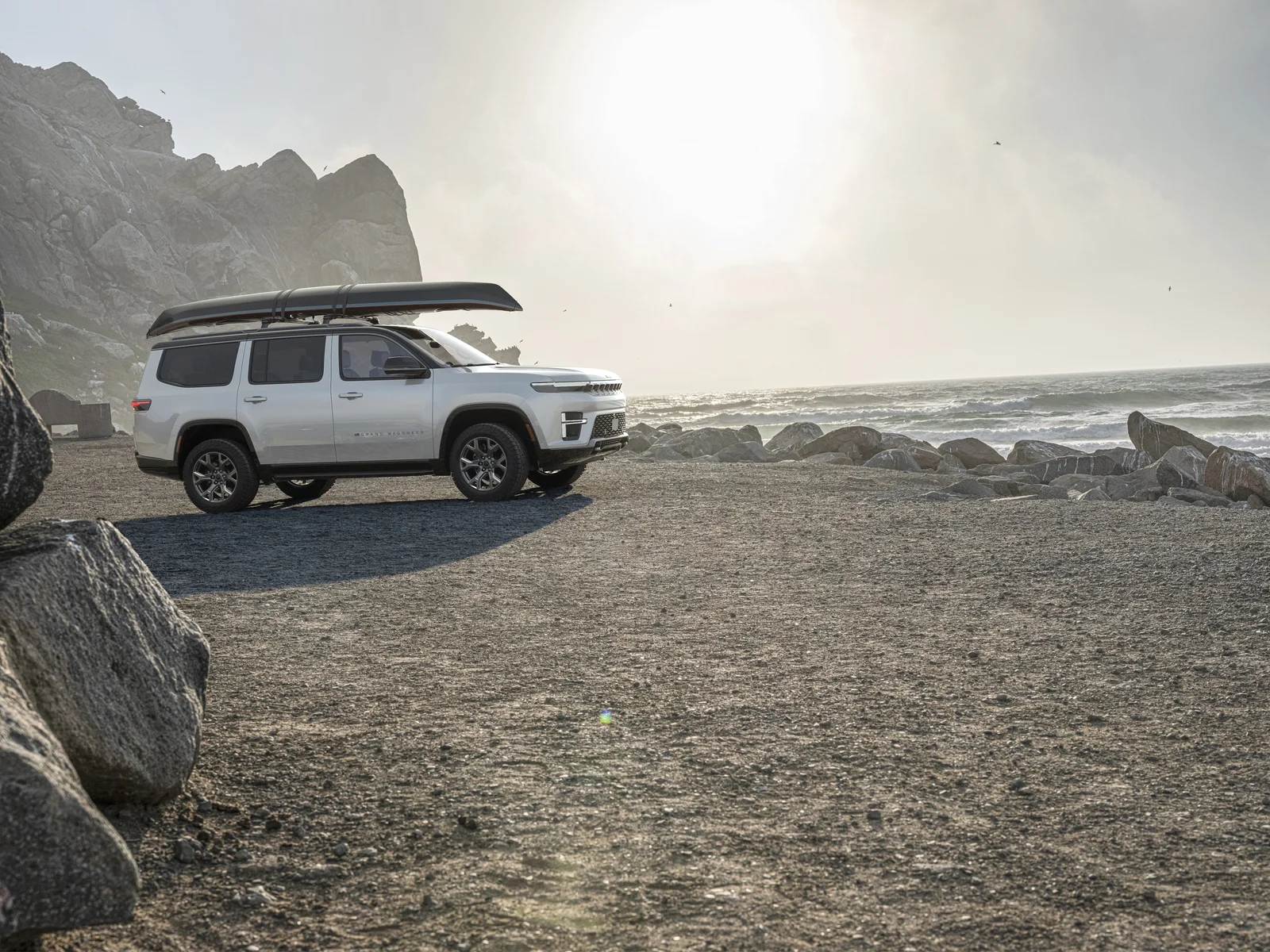 White Jeep Grand Wagoneer with a canoe on the roof parked on a rocky shoreline