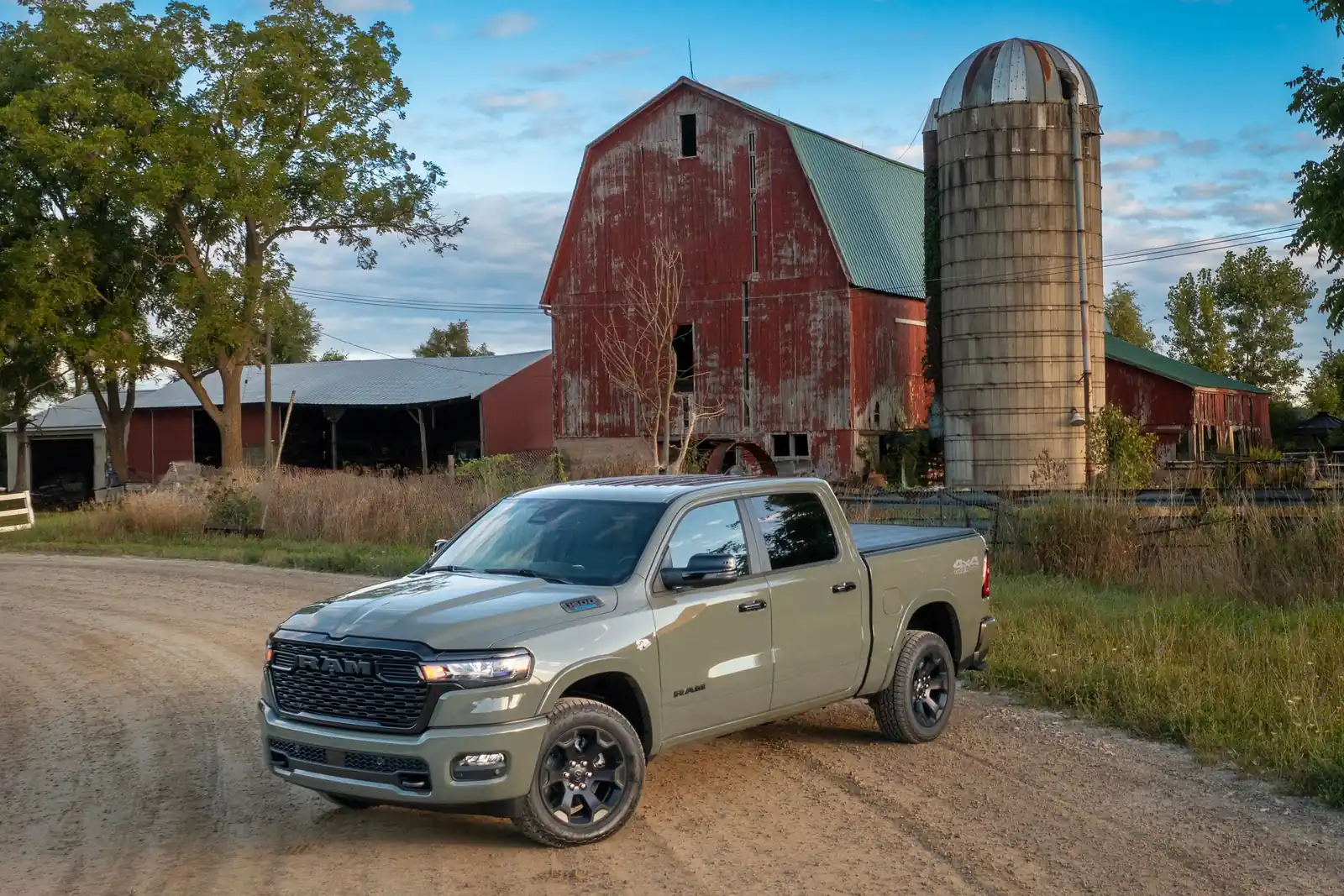 green 2026 Ram 1500 Big Horn crew cab on a gravel farm lane with a red barn and grain silo behind