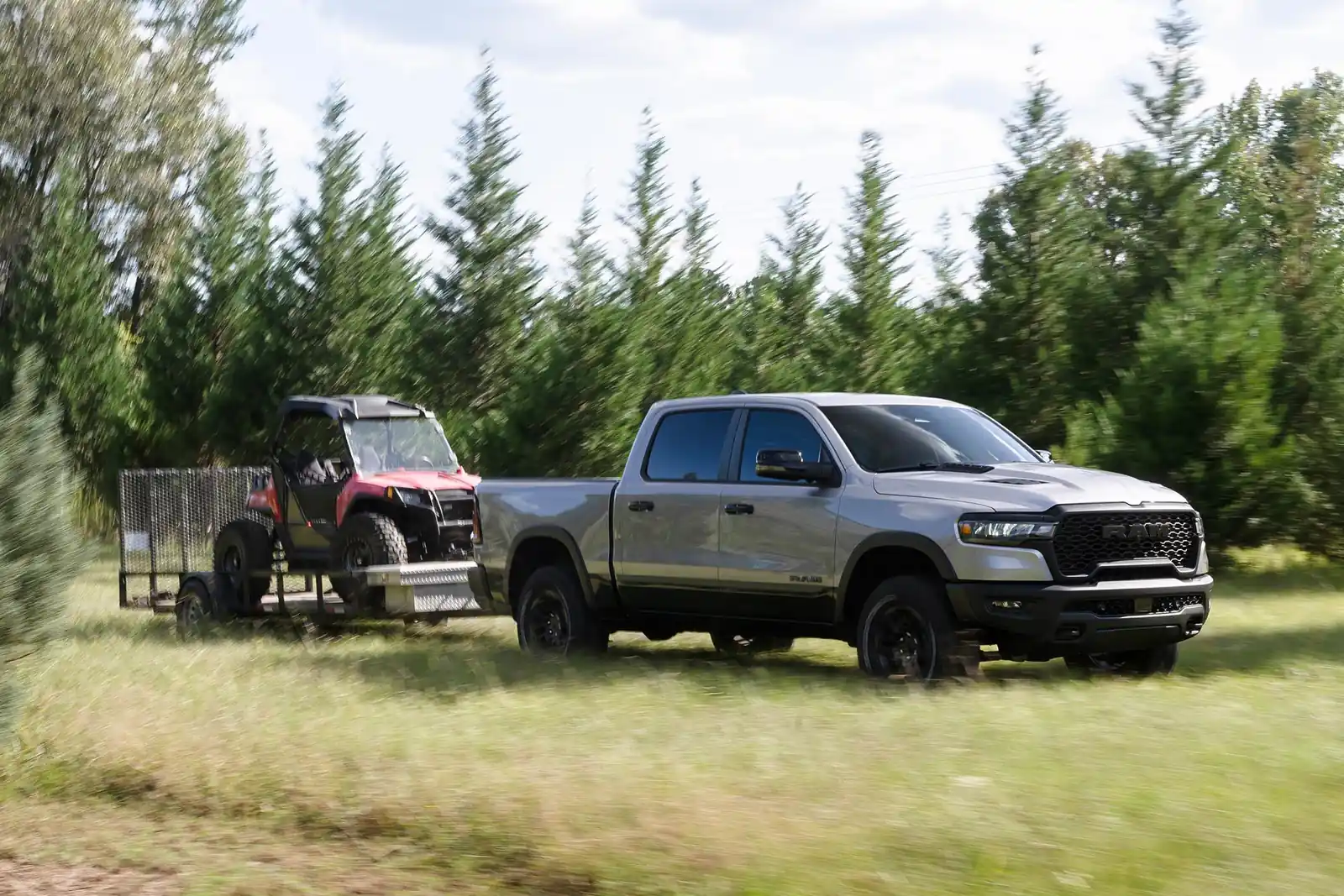 grey Ram 1500 with blacked out grille towing a side by side on a utility trailer past a treeline