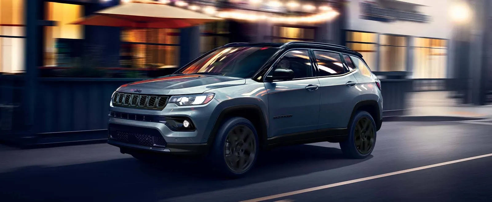 2026 Jeep Compass in gray shown front three quarter driving a city street at dusk