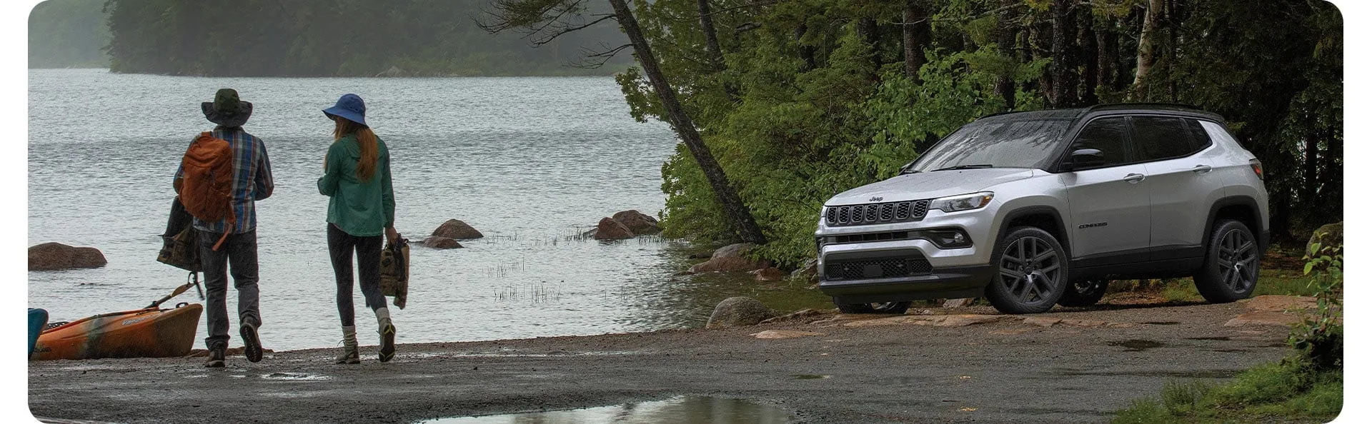 White Jeep Compass parked on a wet gravel lakeshore beside kayaks and trees