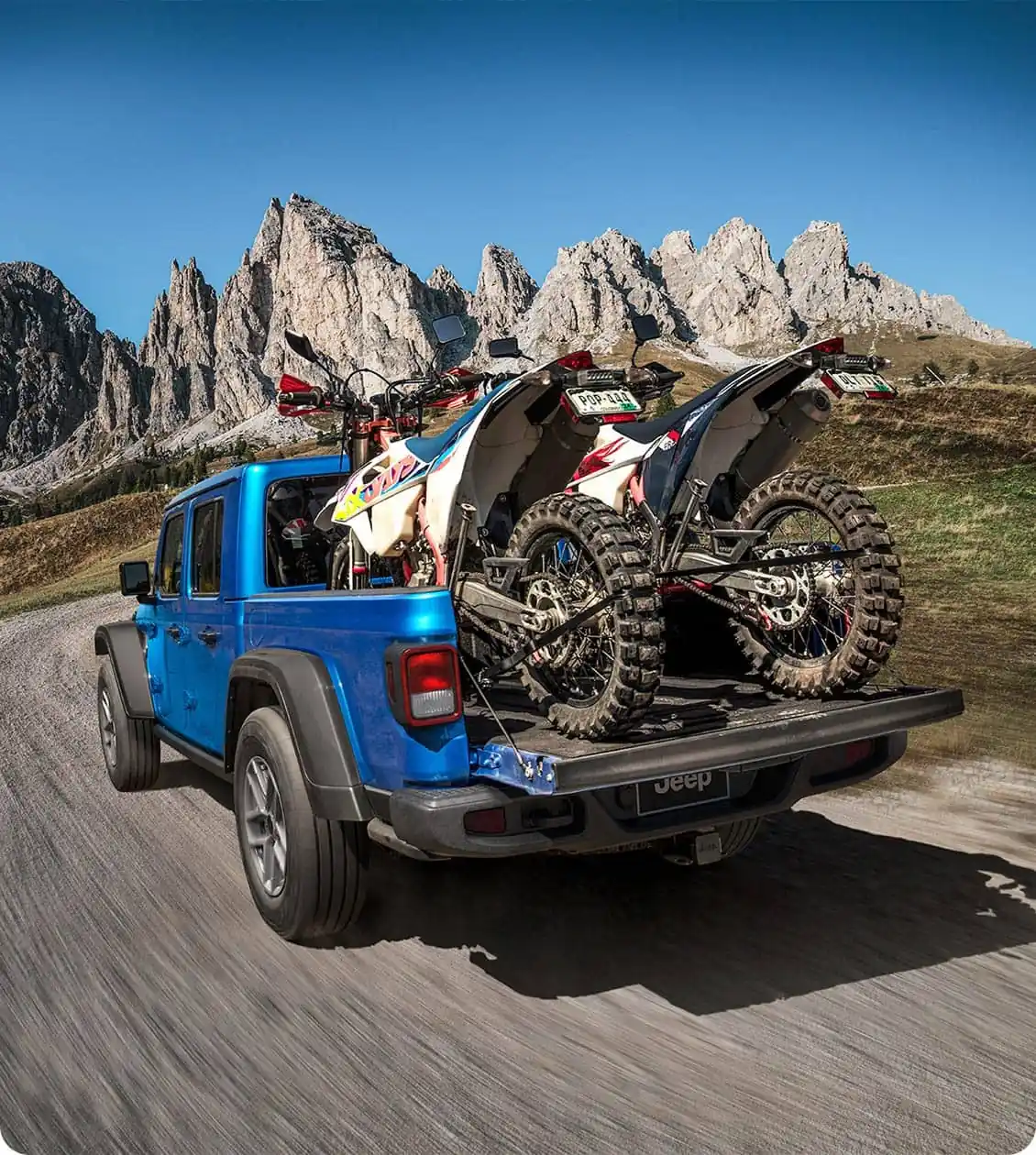 Blue Jeep Gladiator on a gravel road with two dirt bikes strapped in the bed