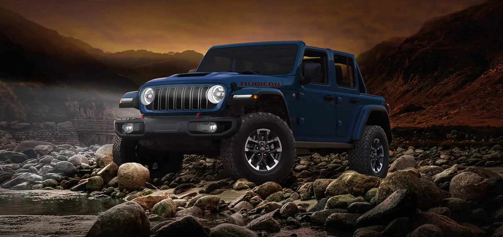 Blue Jeep Wrangler four door with a black hardtop parked on river rocks at dusk