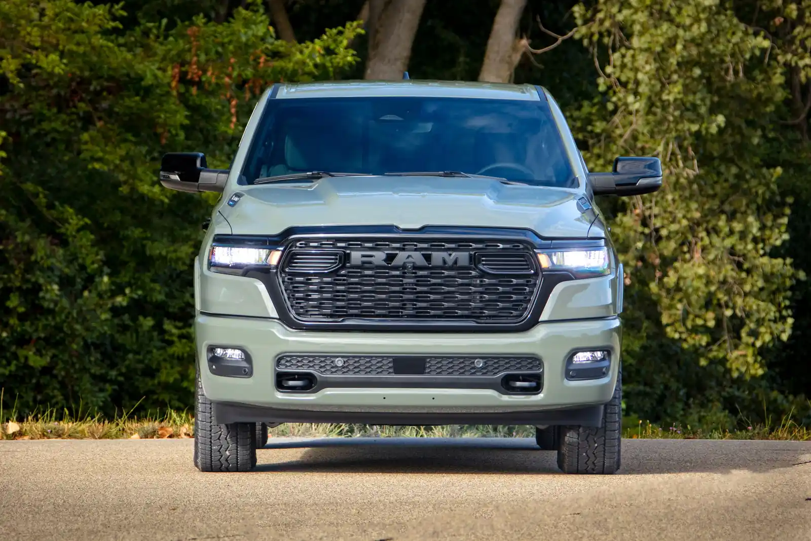 Green Ram 1500 Big Horn crew cab front view on a paved rural lane with a dense treeline