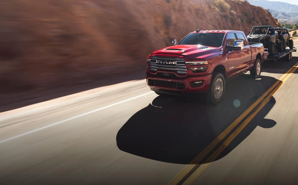 Ram 2500 Laramie in red towing an open trailer loaded with a side by side on the highway