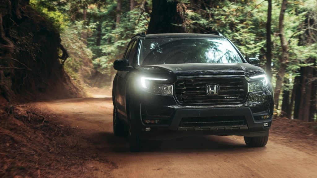 A Honda Passport drives through a forest on a dirt trail, headlights on.
