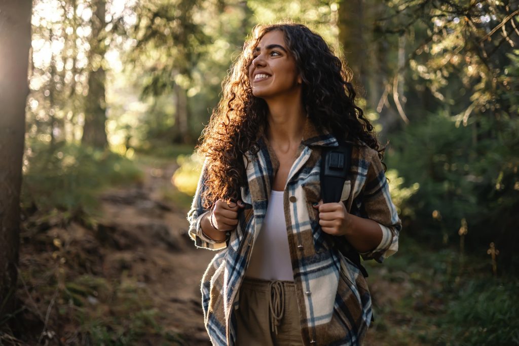 Young woman with curly hair, wearing a backpack, smiles cheerfully while walking through a forest path, feeling the beauty, freedom, and tranquility of nature.