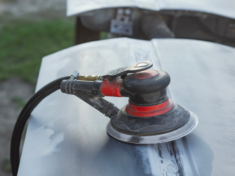 A red and black orbital sander rests on a smooth metal surface, possibly a car hood. The background shows blurred greenery, indicating an outdoor setting.