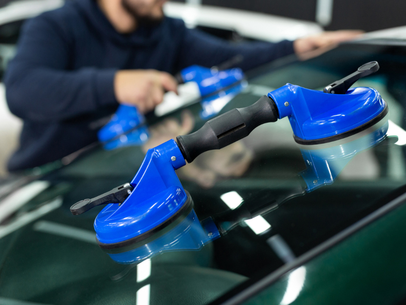 A person in a blue hoodie uses a blue suction cup tool to lift or adjust a car windshield in a workshop. The setting conveys precision and focus.