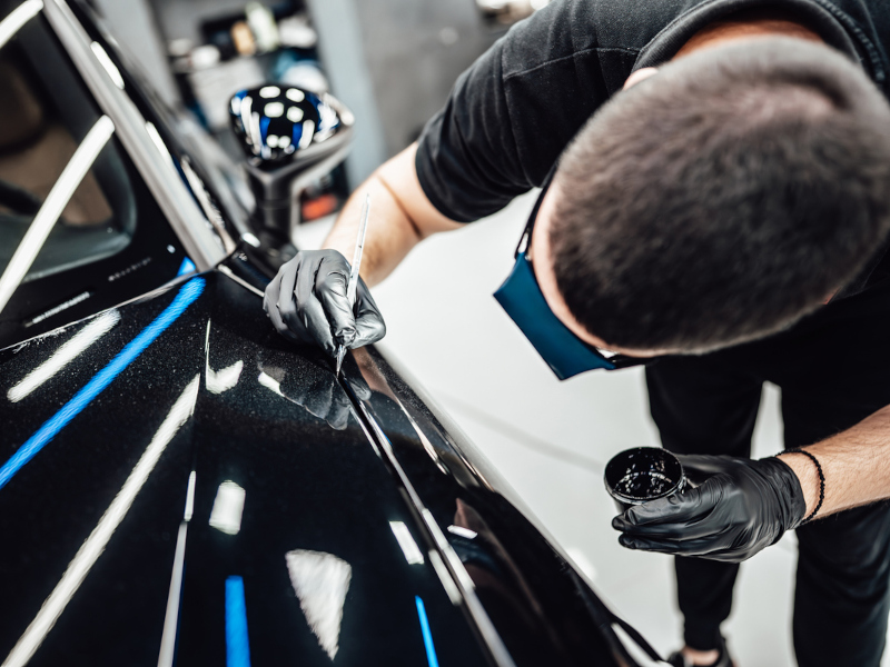 A man in gloves carefully applies black paint to a scratch on a car. The shiny surface reflects bright lights, conveying precision and focus.