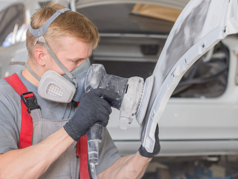 A worker in protective gear sands a car bumper with a power tool. Dust fills the air in the workshop as they focus intently on their task.