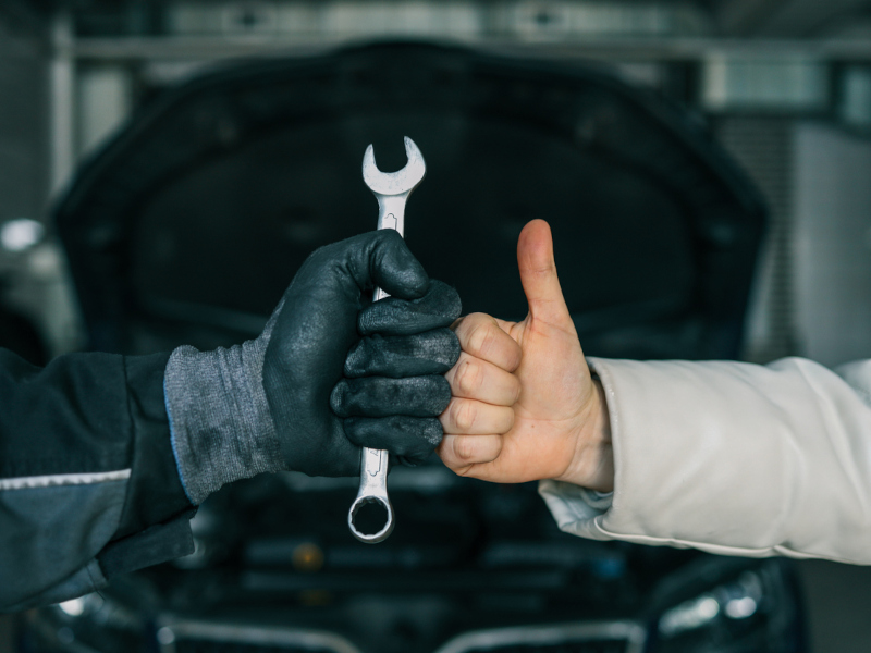 A mechanic's gloved hand holds a wrench, fist-bumping a customer's bare hand with a thumbs-up, in front of an open car hood, symbolizing teamwork.