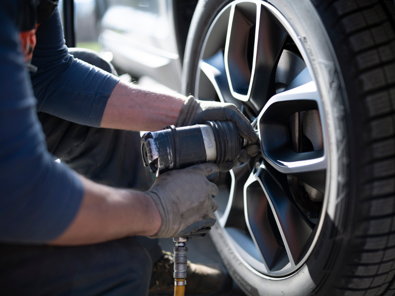 A mechanic uses a pneumatic wrench to tighten the bolts of a car's wheel. The focus is on the wheel and tool, conveying precision and expertise.