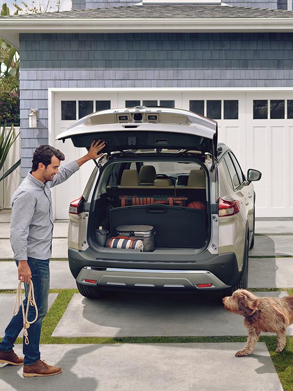 A man loading his dog into the back of his silver Nissan Rogue parked in the driveway.