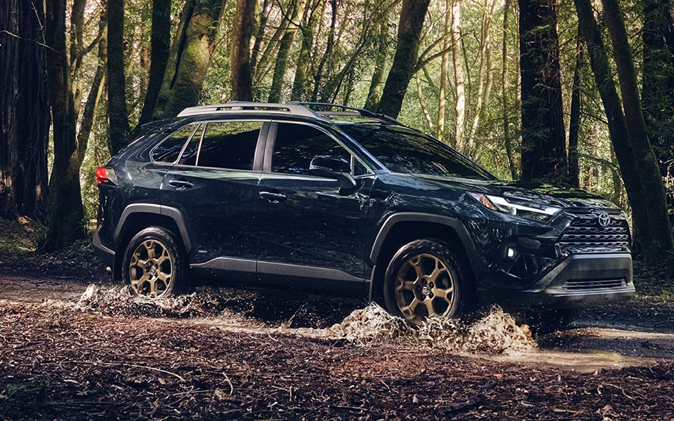 A rugged Toyota RAV4 driving through a forest trail, splashing water as it navigates the terrain. Surrounding trees create a serene, adventurous atmosphere.