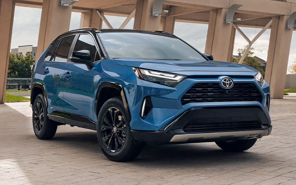 A blue Toyota RAV4 parked in an urban setting under a modern architectural structure. The SUV’s sporty design and black accents stand out.